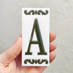 Op de afbeelding: Een rechthoekige witte tegel met de letter "A" in donkergroen. De letter staat in het midden van de tegel en heeft een decoratieve donkergroene rand. Sierlijke donkergroene krullen sieren de boven- en onderkant van de tegel.