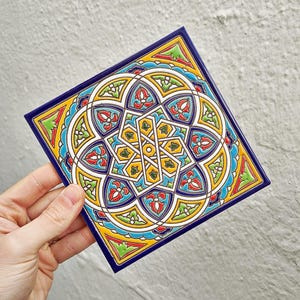 Puede incluir: Azulejo cerámico cuadrado con un detallado patrón geométrico en amarillo, azul, rojo y verde. El azulejo tiene un borde azul oscuro y un acabado brillante. El diseño presenta una forma de estrella central rodeada de elementos florales y geométricos.