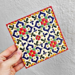 May include: Square ceramic tile with a vibrant floral pattern. The design features red, blue, yellow, and green elements on a cream background. The tile's edges are outlined in red, and the overall aesthetic is reminiscent of traditional decorative tiles.