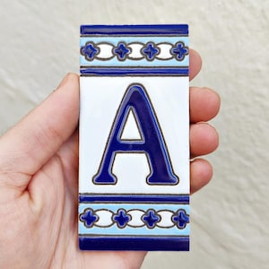 Puede incluir: Azulejo cerámico rectangular con la letra "A" en azul oscuro brillante. La letra está centrada sobre un fondo blanco. Los bordes superior e inferior tienen un diseño decorativo en azul y celeste.