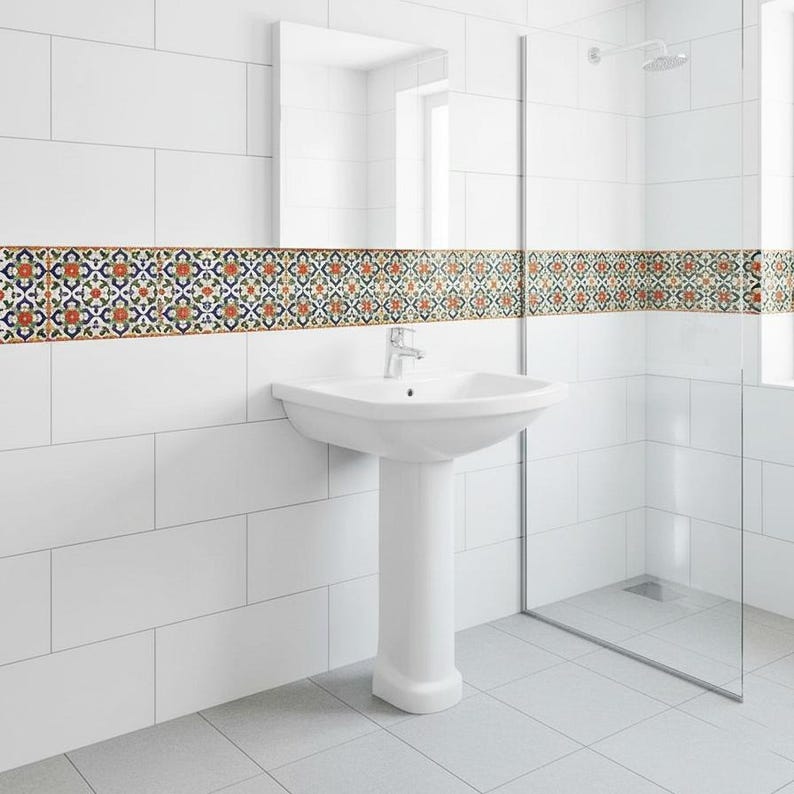 May include: A white pedestal sink with a chrome faucet is centered in a bathroom. The walls are white tile, with a decorative border of patterned tiles in orange, blue, and green. A glass shower enclosure is to the right.