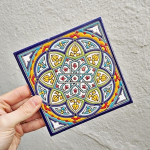 Op de afbeelding: Vierkante keramische tegel met een levendig geometrisch ontwerp. De tegel heeft een centraal sterpatroon omgeven door bloemen en geometrische vormen in rood, geel, groen en blauw, met een donkerblauwe rand. De tegel wordt door een hand tegen een grijze achtergrond gehouden.
