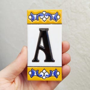 Könnte beinhalten: Rechteckige Keramikfliese mit dem schwarzen Buchstaben "A" auf weißem Hintergrund. Die Fliese hat einen gelb-blauen Dekorrand. Die Fliese wird in einer Hand gehalten.