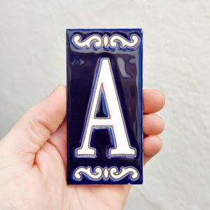 May include: A navy blue rectangular tile with the white letter "A". The letter is centered and framed by a decorative white border. The tile has a glossy finish. The tile is held in a hand.