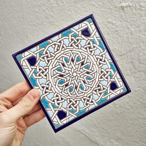 Può includere: Piastrella quadrata in ceramica con un motivo geometrico bianco, azzurro e blu scuro. La piastrella ha un bordo blu scuro ed è tenuta in mano. Il design presenta linee e forme intrecciate.