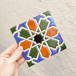 Puede incluir: Azulejo cerámico cuadrado con un patrón geométrico. El diseño presenta una estrella negra central rodeada de formas naranjas, verdes y azules, delineadas en blanco. El azulejo se sostiene sobre un fondo neutro.