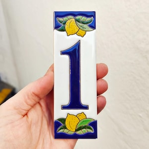 Może przedstawiać: Prostokątny ceramiczny numer domu jeden. Numer jest ciemnoniebieski na białym tle. Górna i dolna część płytki mają niebieską obwódkę z żółtymi cytrynami i zielonymi liśćmi.