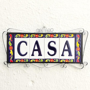 以下が含まれることがあります： 白いタイルに濃い青色の文字で「CASA」と書かれた装飾的な壁掛けサイン。 タイルは、装飾的な渦巻き模様のある黒い錬鉄製のフレームに収められています。 タイルには、花の模様が入ったカラフルなボーダーがあります。