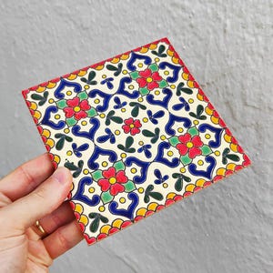 May include: Square ceramic tile with a colorful floral pattern. The design features red flowers, blue swirls, green leaves, and yellow accents on a cream background. The tile has a scalloped red border.