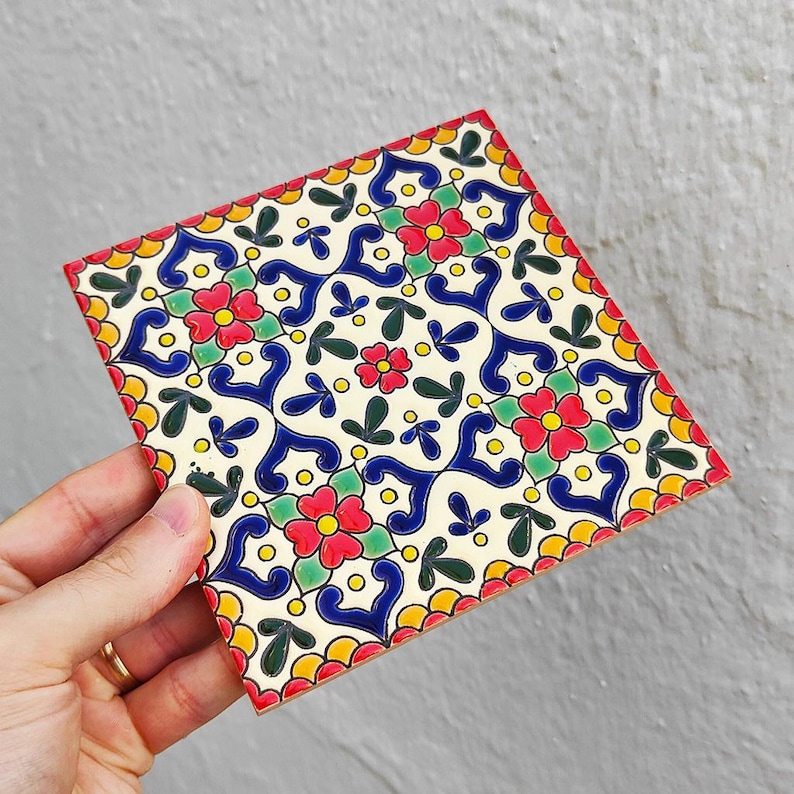 May include: Square ceramic tile with a colorful floral pattern. The design features red flowers, blue swirls, green leaves, and yellow accents on a cream background. The tile has a scalloped red border.