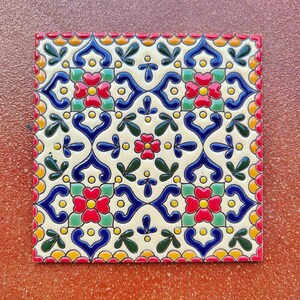 May include: Square ceramic tile with a repeating floral pattern in red, blue, green, and yellow on a cream background. The tile has a red border and is likely for home decor or a craft project.