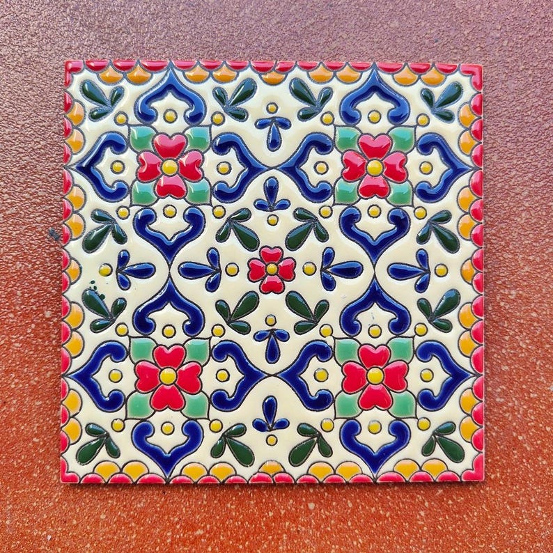 May include: Square ceramic tile with a repeating floral pattern in red, blue, green, and yellow on a cream background. The tile has a red border and is likely for home decor or a craft project.