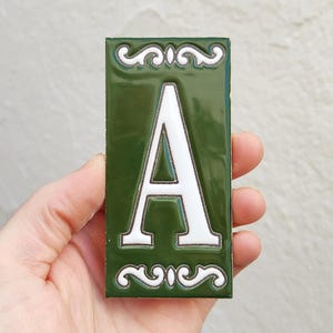 Op de afbeelding: Groene rechthoekige tegel met een witte letter "A" en decoratief krulwerk. De tegel wordt in een hand gehouden, waarbij de glanzende afwerking en contrasterende kleuren worden benadrukt. Een decoratief item voor de interieurdecoratie.