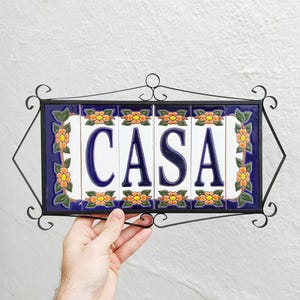 Könnte beinhalten: Dekoratives Schild mit dem Wort "CASA" in dunkelblauen Buchstaben auf weißen Fliesen. Die Fliesen sind in einem schwarzen Schmiedeeisenrahmen mit floralen und Blattakzenten eingefasst. Der Rand ist dunkelblau mit orangefarbenen Blumen.