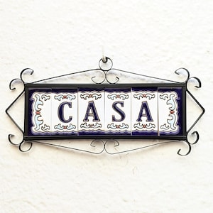 Pode incluir: Uma placa decorativa com a palavra "CASA" em letras azuis sobre azulejos brancos, emoldurada por uma borda de ferro forjado preto. Os azulejos têm uma borda azul com padrões decorativos. A placa está pendurada numa parede branca.