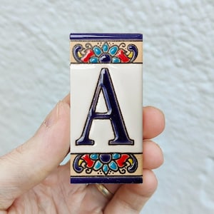 Puede incluir: Una baldosa cerámica rectangular con la letra "A" en azul marino sobre fondo blanco. La baldosa está bordeada con un ribete azul marino y patrones florales decorativos en rojo, azul y amarillo.