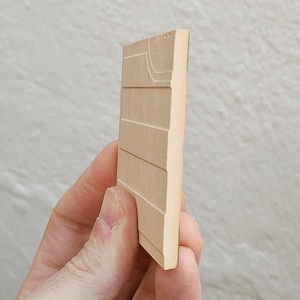 May include: A light brown, rectangular wooden block with horizontal and curved carved lines. The block is held in a hand, showcasing the side profile and texture of the wood. The carving appears to be a map or architectural detail.