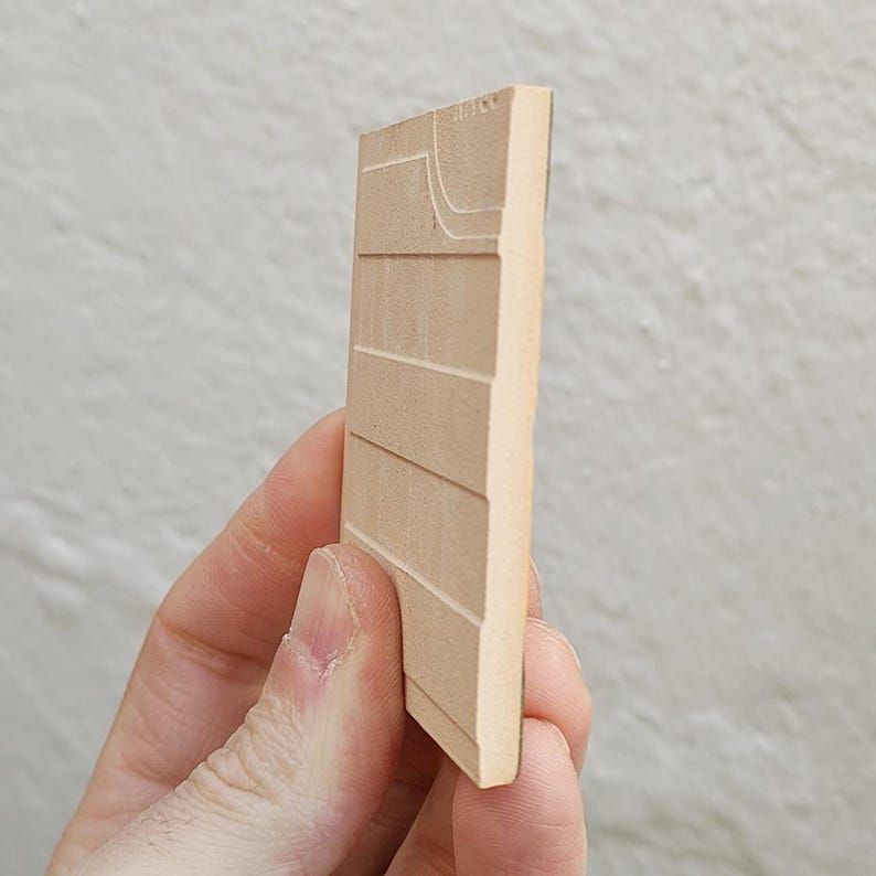 May include: A light brown, rectangular wooden block with horizontal and curved carved lines. The block is held in a hand, showcasing the side profile and texture of the wood. The carving appears to be a map or architectural detail.