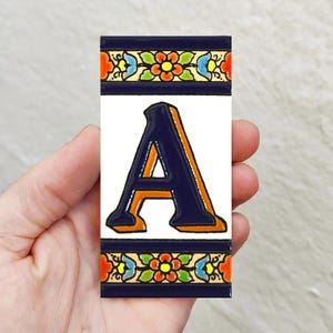 Puede incluir: Azulejo decorativo con la letra "A" en azul marino con contorno naranja. El azulejo tiene un fondo blanco y está enmarcado por un borde azul marino con motivos florales coloridos. El azulejo es rectangular.