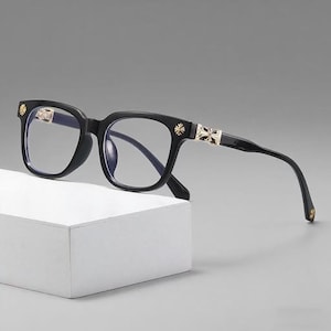 Puede incluir: Gafas negras con montura cuadrada. Las patillas y las esquinas presentan detalles decorativos dorados. Las lentes son transparentes. Las gafas están sobre una superficie blanca.