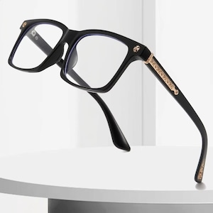 May include: Black eyeglasses with a square frame design. The glasses have gold-colored accents on the temples and frame corners. The lenses are clear, and the arms curve down to the ear pieces. The glasses are set against a white background.