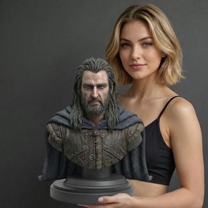Pode incluir: Um busto esculpido de um homem barbudo com cabelo comprido, vestindo uma peça de peito de couro castanho texturizado com tachas metálicas e uma capa cinzenta escura. A figura está montada numa base de pedestal circular preta.