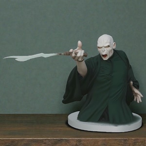 Puede incluir: Busto de un personaje con rostro pálido, boca abierta y túnicas verde oscuro, sosteniendo una varita con un efecto de energía blanca. La figura está sobre una base circular blanca, sobre un fondo verde apagado. La varita es marrón.