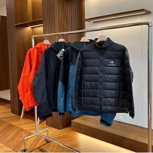 May include: A metal clothing rail holds a selection of jackets. There's a bright orange hooded jacket, a black jacket, a black and blue jacket, and a black quilted jacket. The jackets hang against a backdrop of wooden panelling and flooring.