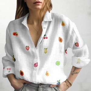 Könnte beinhalten: Weißes Button-Down-Hemd mit langen Ärmeln, verziert mit farbenfrohen Aquarell-Fruchtillustrationen. Das Hemd zeigt eine Vielzahl von Früchten, darunter Erdbeeren, Zitronen und Pfirsiche. Das Hemd ist aus leichtem Stoff gefertigt.