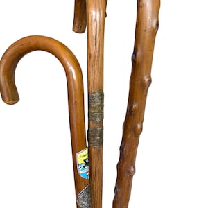 Op de afbeelding: Drie houten wandelstokken met gebogen handvatten. De stokken hebben een natuurlijke houtnerf, knoesten en diverse metalen badges en kleurrijke stickers op de schachten. De badges bevatten gegraveerde teksten en wapenschilden.