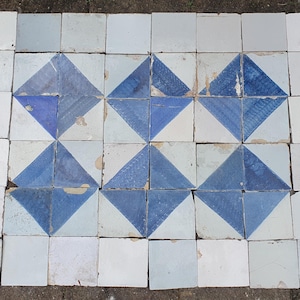 Puede incluir: Una colección de baldosas de cerámica cuadradas dispuestas en una cuadrícula rectangular. Las baldosas presentan un patrón geométrico creado alternando baldosas de color blanco roto con baldosas pintadas con triángulos azules y blancos.