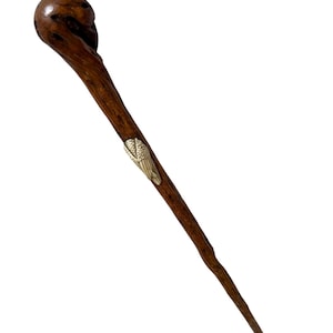 Könnte beinhalten: Ein langer, schlanker Haarpick aus dunkelbraunem Holz mit einem abgerundeten, strukturierten Kopf. Der Schaft ist mit einer kleinen, metallischen, goldfarbenen Verzierung in Form eines stilisierten Flügels oder Blattes versehen.