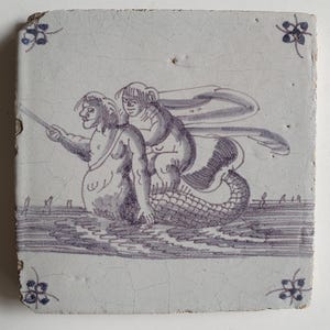 Op de afbeelding: Vierkante keramische tegel met een handgeschilderde illustratie in blauw- en wittinten. De afbeelding toont een zeemeerminachtige figuur met twee figuren op haar rug, die een lange stok vasthouden. Bloemenaccenten bevinden zich in elke hoek.