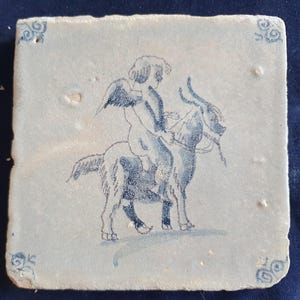 Puede incluir: Un azulejo de cerámica cuadrado con una superficie de color beige claro que presenta una ilustración en tinta azul de una figura alada montada sobre una cabra. El azulejo tiene patrones decorativos de volutas azules en las cuatro esquinas.