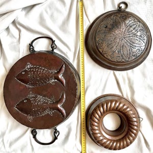 Op de afbeelding: Drie koperen keukenmallen, waaronder een ronde pan met twee reliëfvissen, een ronde vergiet met een geperforeerd bloemmotief en een geribbelde ringvorm. Een geel meetlint toont een lengte van 25 tot 65 cm.
