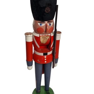 Op de afbeelding: Een houten notenkraker soldatenfiguur. De figuur is beschilderd met een rode jas, een blauwe broek en een zwarte hoed. Het heeft een geschilderd gezicht met blauwe ogen en houdt een houten geweer vast. De basis is groen.