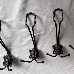 Op de afbeelding: Vier zwarte metalen wandhaken op een rij. Elke haak heeft een decoratieve, sierlijke basis met drie montagegaten en een lange, slanke verticale arm die aan de bovenkant een lus vormt, aangevuld met twee kleinere, gebogen haken aan de zijkanten.