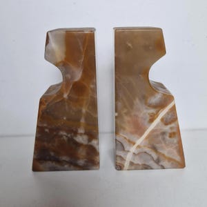 May include: A pair of decorative bookends. Each bookend is made of a marbled stone with shades of brown, tan, and white. The bookends have a unique, asymmetrical design with a curved cutout near the top.
