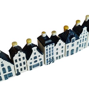 May include: A collection of miniature ceramic houses. The houses have white facades, dark roofs, and blue window and door details. Each house has a unique shape and design, with yellow chimney tops. The houses are arranged in a diagonal line.