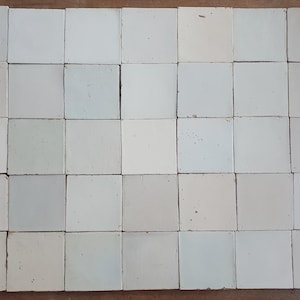 Puede incluir: Una colección de baldosas cuadradas de cerámica blanquecina dispuestas en un patrón de cuadrícula. Las baldosas tienen una superficie ligeramente irregular y varían en tonalidad, desde crema claro hasta gris pálido. El efecto general es una apariencia rústica y texturizada.