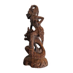 May include: A detailed, hand-carved wooden sculpture depicting two figures. One figure is atop another, with intricate details in their clothing and facial expressions. The sculpture is a warm brown color and stands on a carved base.