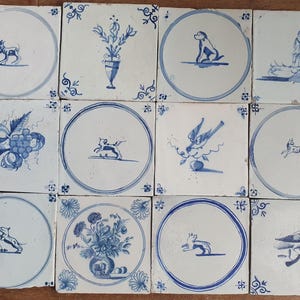 May include: A collection of twelve square ceramic tiles featuring blue and white hand-painted designs. The tiles display various motifs including animals, floral arrangements in vases, fruit clusters, and human figures, all set against a white background.