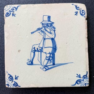 May include: Square, off-white ceramic tile featuring a blue illustration of a person playing a flute. The figure is seated and wearing a hat and period clothing. Blue decorative corner accents. The tile has a slightly aged appearance.