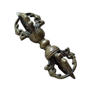 Op de afbeelding: Een bronzen vajra, een ritueel object met sierlijke details. De handgreep heeft een bolvormige vorm en de uiteinden hebben een gestileerd ontwerp met gebogen elementen. Het algehele ontwerp suggereert een religieus of ceremonieel doel.