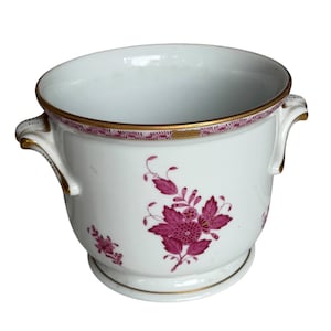 May include: A white porcelain cachepot featuring a magenta floral pattern on the exterior. The vessel has two side handles, a gold-trimmed rim, and a gold-accented base. The design includes a central magenta flower motif with leaves.