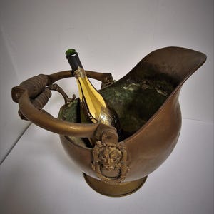 Op de afbeelding: Een vintage bronzen champagne emmer met een fles erin. De emmer heeft een handvat en een decoratief gezicht aan de zijkant. De fles heeft een gouden label en een groene dop.
