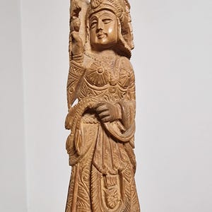 Può includere: Intagliatura in legno dettagliata di una figura in abito tradizionale, con motivi intricati e un'espressione serena. La figura è adornata da un copricapo e tiene in mano un oggetto, mostrando l'abilità artigianale. La scultura è di colore marrone chiaro.