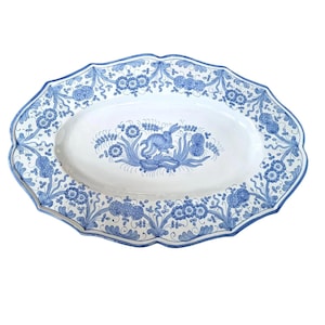 Op de afbeelding: Ovale keramische serveerschaal met een geschulpte rand. De witte schaal heeft een blauwe bloemenrand en een centraal ontwerp van een blauw konijn omringd door bloemen en bladeren. Een decoratief stuk om eten te serveren.