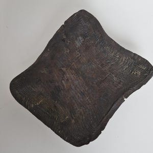 Op de afbeelding: Een donkerbruin, vierkant houten object met een getextureerd oppervlak. De houtnerf is zichtbaar en de randen zijn licht afgerond. Het object lijkt met de hand gesneden of natuurlijk gevormd, met een rustieke uitstraling.