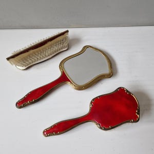 Könnte beinhalten: Ein Vintage-Kosmetikset mit einer Haarbürste und zwei Handspiegeln. Die Spiegel und die Bürste haben rote Emaille-Griffe und -Rahmen mit goldfarbenen Zierleisten. Die Bürste hat weiße Borsten.